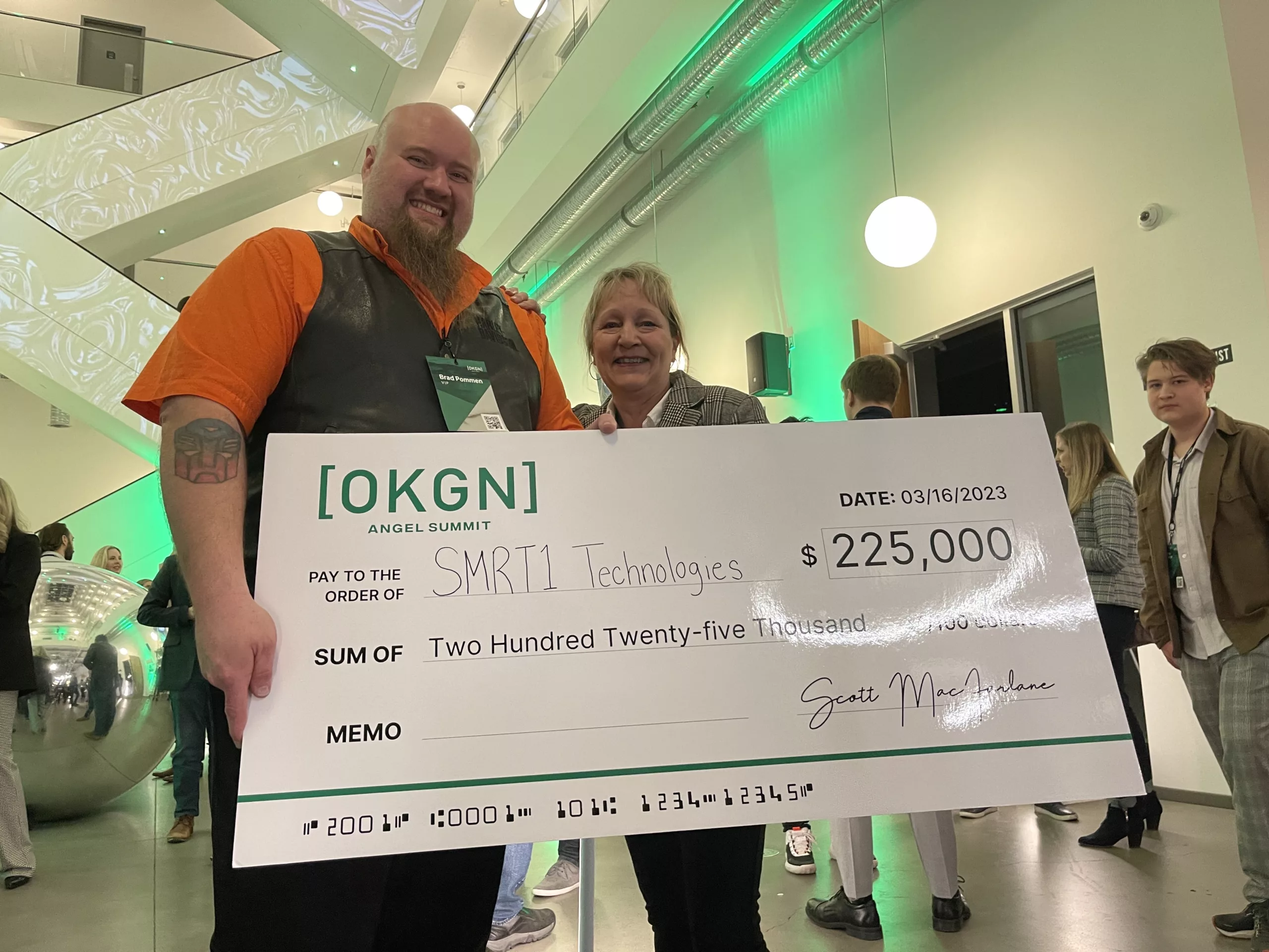 Brad Pommen CEO SMRT1 Technologies standing beside Margret Jennings COO SMRT1 Technologies holding large cheque for $225,000 for first place in OKGN Angel Summit. competition. They are smiling.